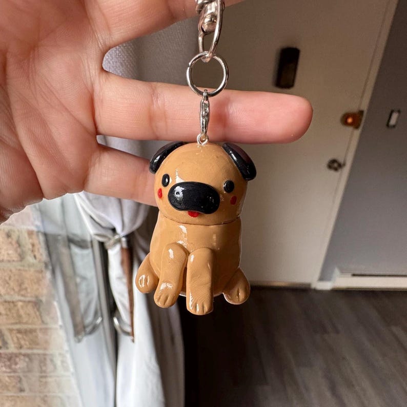 May include: A tan pug keychain with black accents on the nose and ears. The pug has red cheeks and a red mouth. The keychain is attached to a silver metal ring and clasp.