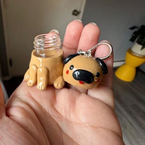 May include: A handmade, tan-colored miniature bottle with a clear glass top and a pug-shaped keychain. The pug keychain has black ears, a black nose, and red accents. The bottle is designed to look like a pug is holding it.