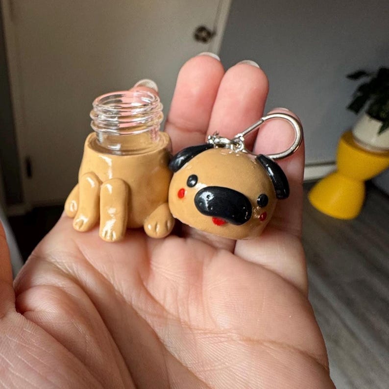 May include: A handmade, tan-colored miniature bottle with a clear glass top and a pug-shaped keychain. The pug keychain has black ears, a black nose, and red accents. The bottle is designed to look like a pug is holding it.