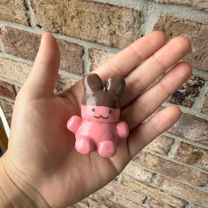 May include: A small, glossy, hand-sculpted clay figurine shaped like a seated bear. The bear has a dark brown head and ears, with a light pink body, arms, and legs. A simple, hand-painted black line forms a small, minimalist face with eyes and a mouth.