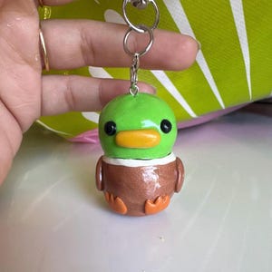 以下が含まれることがあります： 小さな漫画風のアヒルの手作りキーホルダー。アヒルは緑色の頭、オレンジ色のくちばし、オレンジ色の足を持つ茶色の体を持っています。銀色の金属製の留め金が付いています。