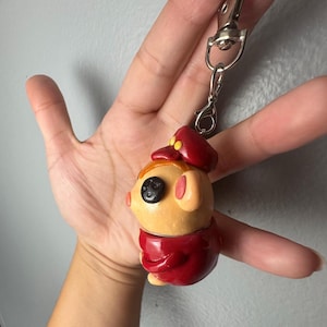 Op de afbeelding: Een kleine, gebeeldhouwde sleutelhangerfiguur met een beige, afgerond personage met &eacute;&eacute;n zwart knoopoog, gekleed in een donkerrode pet en een bijpassende donkerrode jas. De figuur is bevestigd aan een zilverkleurige metalen karabijnhaak en sleutelring.