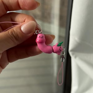 May include: A pink charm featuring two connected figures with simple black facial features. One figure is elongated and curved, while the other is round with a small green sprout on top. The charm is attached to a thin pink cord with silver metal hardware.