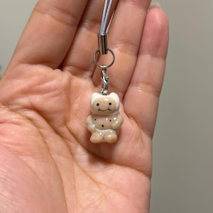 May include: A small, light brown and white ceramic bear charm attached to a silver-toned metal lobster clasp and a light blue nylon cord. The bear figure features a smooth, glossy finish with painted black eyes, a simple smile, and small dots.