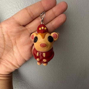 Op de afbeelding: Een polymeerklei sleutelhanger met een beige, beerachtig figuurtje in een donkerrood uniform met bijpassende pet. Het figuurtje heeft zwarte ronde ogen, een kleine roze neus en beige oren. Aan de bovenkant zit een zilverkleurige metalen ring.