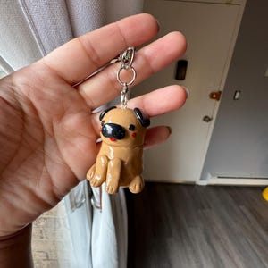 May include: A tan pug keychain with black ears and a black snout. The pug is sitting with its legs forward. The keychain has a silver metal ring and clasp.