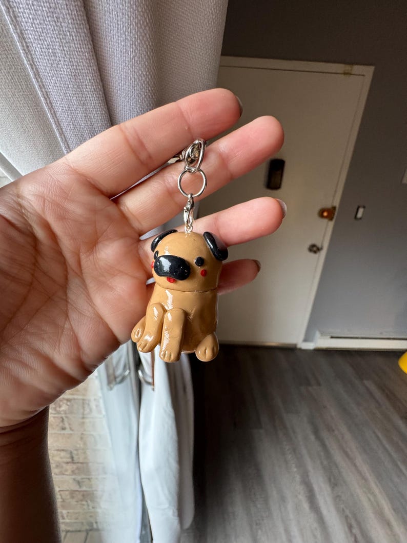 May include: A tan pug keychain with black ears and a black snout. The pug is sitting with its legs forward. The keychain has a silver metal ring and clasp.