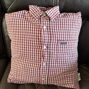 May include: A square cushion made from a red, white, and black plaid button-down shirt. The cushion has a collar, buttons, and a pocket. The cushion is on a brown leather sofa.