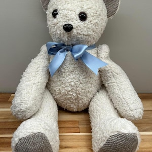 May include: A soft white teddy bear with a light blue ribbon bow. The bear has dark brown eyes, a black nose, and grey fabric on the ears and feet. It is sitting on a wooden surface. The bear is a cuddly toy.