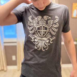Pode incluir: Uma t-shirt de manga curta cinzento-carvão com uma impressão gráfica branca desgastada de uma águia de duas cabeças centrada no peito. O design da águia inclui um escudo com uma cruz e quatro letras cirílicas no centro do emblema.