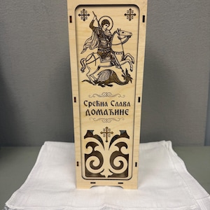 May include: A light-colored wooden box with a carved image of a knight on horseback slaying a dragon. The box has decorative cutouts and text in a foreign language. The box is displayed on a white cloth.