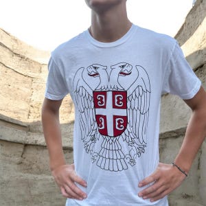 Pode incluir: Uma t-shirt branca de manga curta com uma ilustração de linhas pretas de uma águia de duas cabeças. Centrado no peito da águia está um escudo vermelho com uma cruz branca e quatro caracteres cirílicos em forma de S em cada quadrante.
