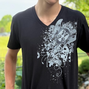 Pode incluir: Camiseta preta com decote em V com um design gráfico detalhado em prata e branco na frente. O design inclui uma caveira, asas e a palavra "SERBIA". A camiseta tem mangas curtas.