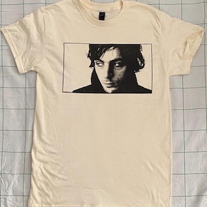 以下が含まれることがあります： クリーム色のTシャツ。長方形のフレームの中に白黒の肖像画が描かれています。肖像画は、暗い髪と物思いにふける表情をした人物を描いています。Tシャツは半袖で、クルーネックです。