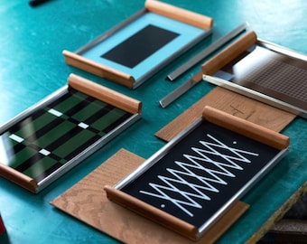 Puede incluir: Varios bandejas rectangulares con diversos diseños y colores se muestran sobre una superficie turquesa. Las bandejas tienen asas de madera y marcos plateados. Los diseños incluyen un rectángulo negro, un estampado de cuadros verdes y negros y un patrón de zigzag blanco sobre fondo negro.