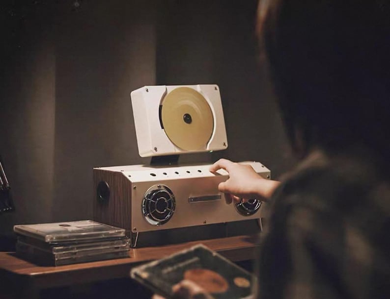 Puede incluir: Un reproductor de CD port&aacute;til blanco y con acabado de madera, con una ranura vertical para discos, dos altavoces frontales circulares con rejillas plateadas y un panel de control superior. Una pila de discos compactos se sit&uacute;a a la izquierda.