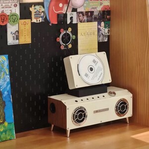 Puede incluir: Un reproductor de CD rectangular de color crema con paneles laterales de imitaci&oacute;n madera y dos altavoces circulares con rejillas plateadas. El dispositivo cuenta con una ranura para CD superior, un mando de volumen plateado y cuatro patas.