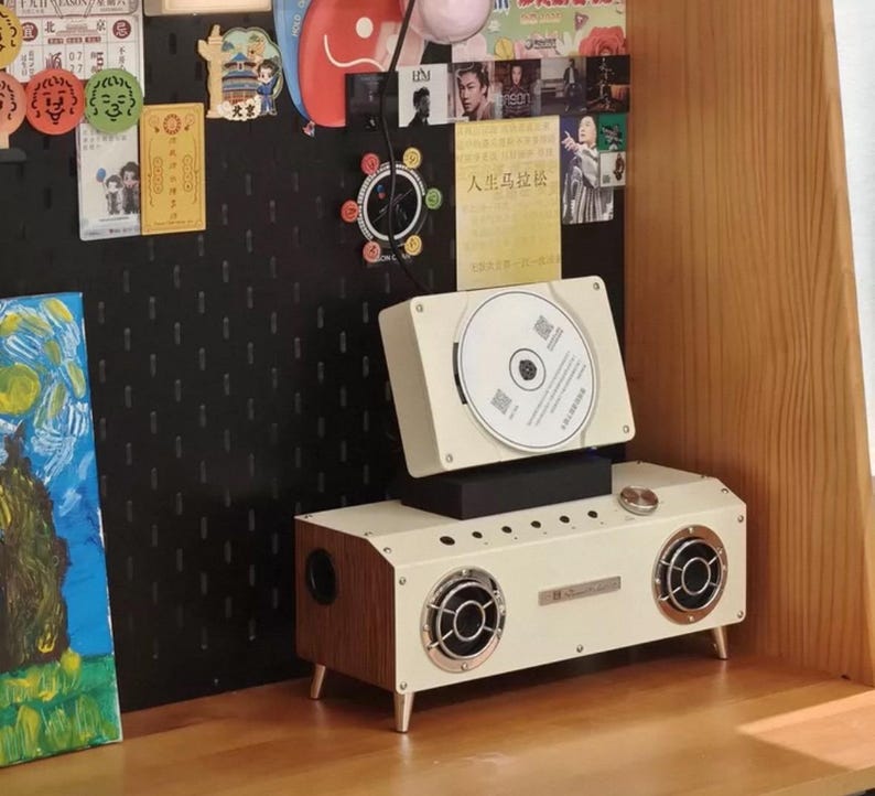 Puede incluir: Un reproductor de CD rectangular de color crema con paneles laterales de imitaci&oacute;n madera y dos altavoces circulares con rejillas plateadas. El dispositivo cuenta con una ranura para CD superior, un mando de volumen plateado y cuatro patas.