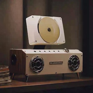 Puede incluir: Un reproductor de audio rectangular blanco con dos altavoces circulares con rejillas plateadas, un reproductor de discos montado en la parte superior con un disco dorado y una perilla de control met&aacute;lica. Tiene paneles laterales de madera.