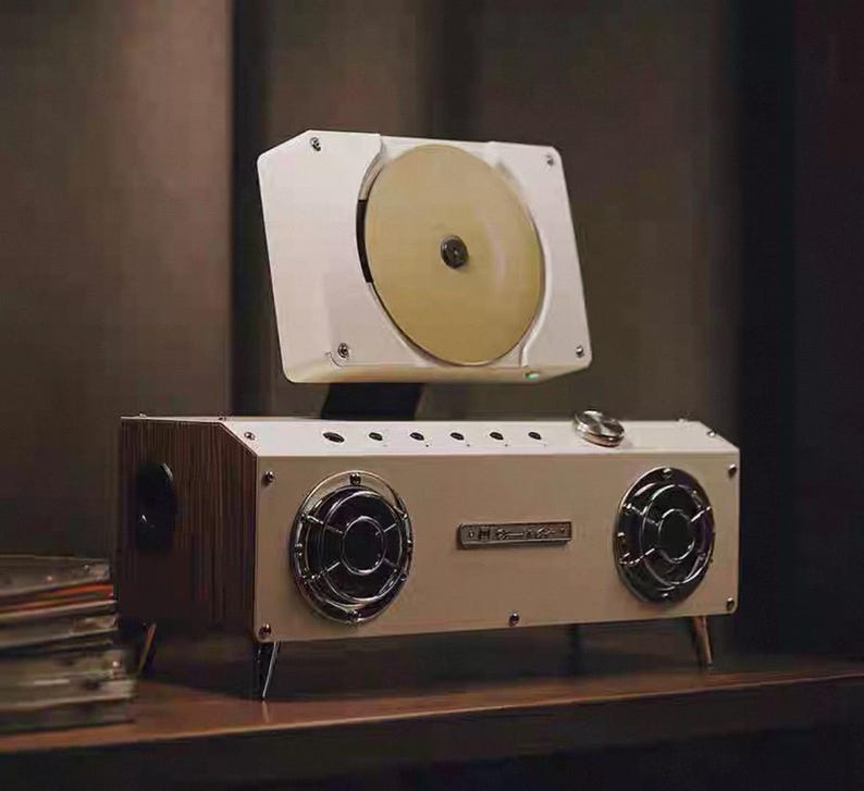 Puede incluir: Un reproductor de audio rectangular blanco con dos altavoces circulares con rejillas plateadas, un reproductor de discos montado en la parte superior con un disco dorado y una perilla de control met&aacute;lica. Tiene paneles laterales de madera.