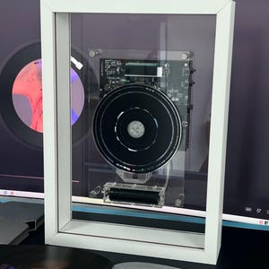 May include: A framed artwork with a black circular component and a circuit board, presented within a white frame. The clear front allows a view of the internal details. The piece has a modern aesthetic.