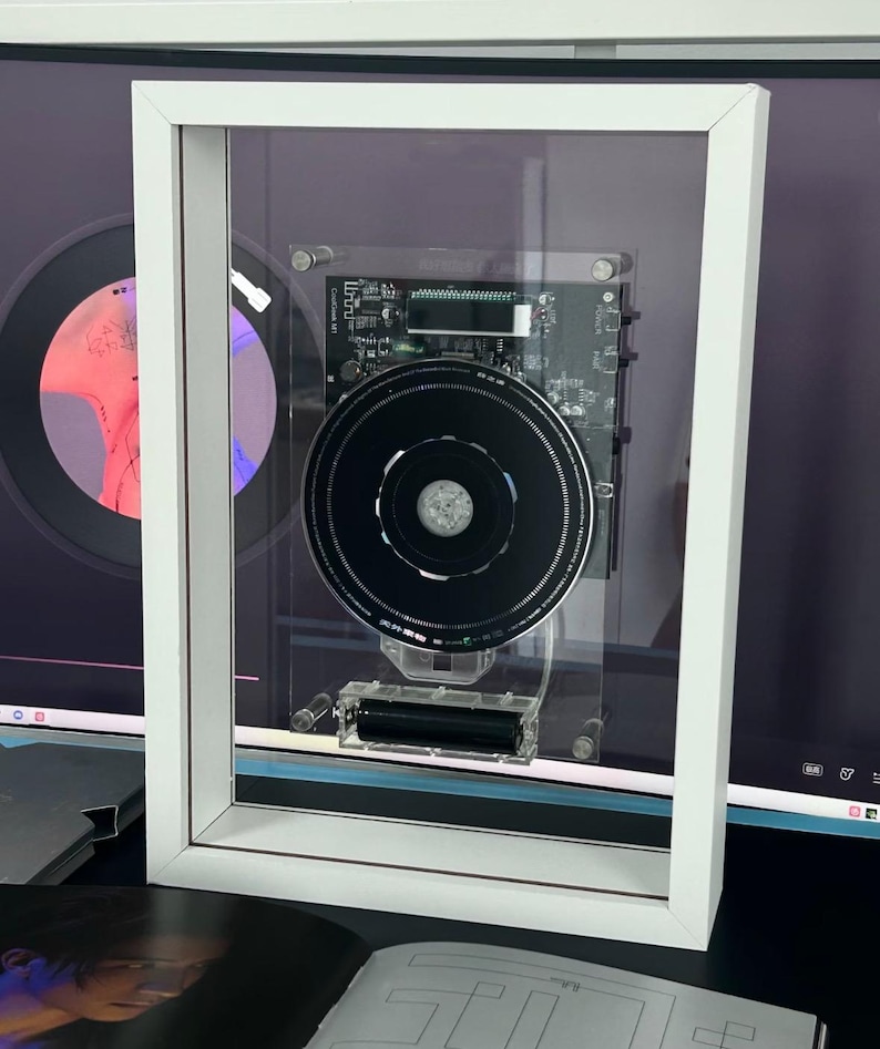 May include: A framed artwork with a black circular component and a circuit board, presented within a white frame. The clear front allows a view of the internal details. The piece has a modern aesthetic.