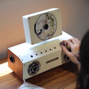 Puede incluir: Un reproductor de CD blanco con vetas de madera, ranura vertical para discos, dos altavoces frontales circulares con rejillas plateadas, una perilla de control superior y una serie de botones circulares peque&ntilde;os.