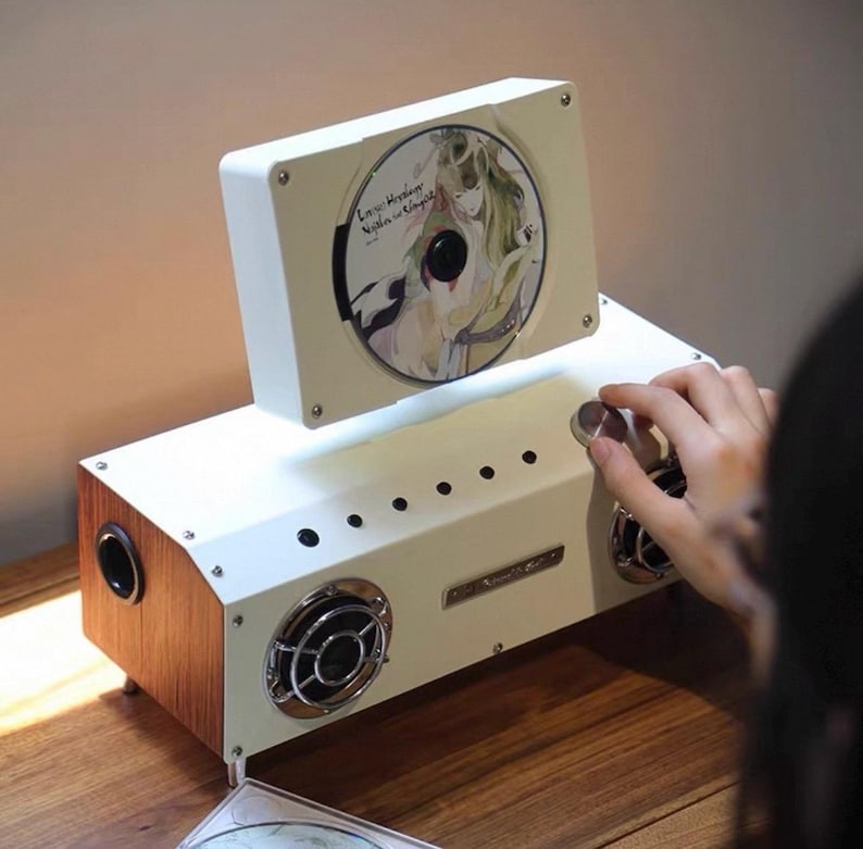 Puede incluir: Un reproductor de CD blanco con vetas de madera, ranura vertical para discos, dos altavoces frontales circulares con rejillas plateadas, una perilla de control superior y una serie de botones circulares peque&ntilde;os.