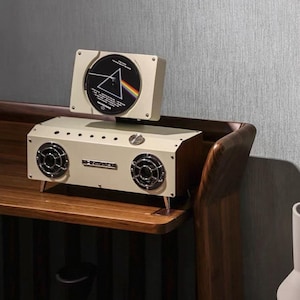 Puede incluir: Un altavoz rectangular de color crema con dos controladores circulares frontales y una ranura vertical superior que sostiene un CD con el gr&aacute;fico del prisma de Pink Floyd Dark Side of the Moon. El dispositivo tiene un mando de control met&aacute;lico.