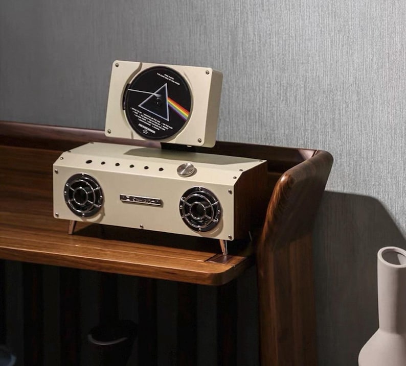 Puede incluir: Un altavoz rectangular de color crema con dos controladores circulares frontales y una ranura vertical superior que sostiene un CD con el gr&aacute;fico del prisma de Pink Floyd Dark Side of the Moon. El dispositivo tiene un mando de control met&aacute;lico.