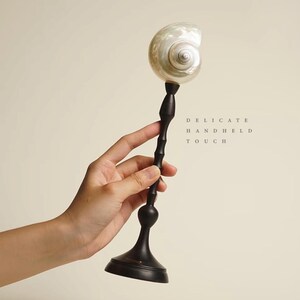 Op de afbeelding: Een decoratief object met een parelmoeren schelp op een donkere, slanke, bamboe-achtige steel en een ronde basis. De tekst "DELICATE HANDHELD TOUCH" staat rechts afgedrukt. Het object wordt in een hand gehouden.