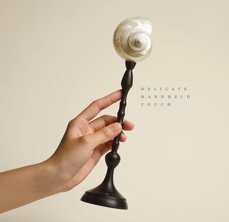 Op de afbeelding: Een decoratief object met een parelmoeren schelp op een donkere, slanke, bamboe-achtige steel en een ronde basis. De tekst "DELICATE HANDHELD TOUCH" staat rechts afgedrukt. Het object wordt in een hand gehouden.