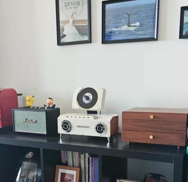 Puede incluir: Un reproductor de CD port&aacute;til blanco y de madera con mecanismo de carga superior y dos altavoces frontales, colocado en un estante negro junto a un altavoz Marshall verde y un cofre de almacenamiento de madera.