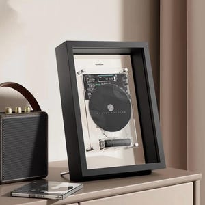 May include: A black framed CD player with a clear front panel, displaying a black CD and circuit board. The frame is angled, resting on a silver stand. A small, black speaker with a brown handle and a CD case are also visible.