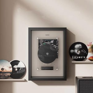 May include: A black framed vinyl record player with a black record and a digital display. Two vinyl records are on a shelf to the left, and two more are on a shelf to the right. The wall is a neutral colour.
