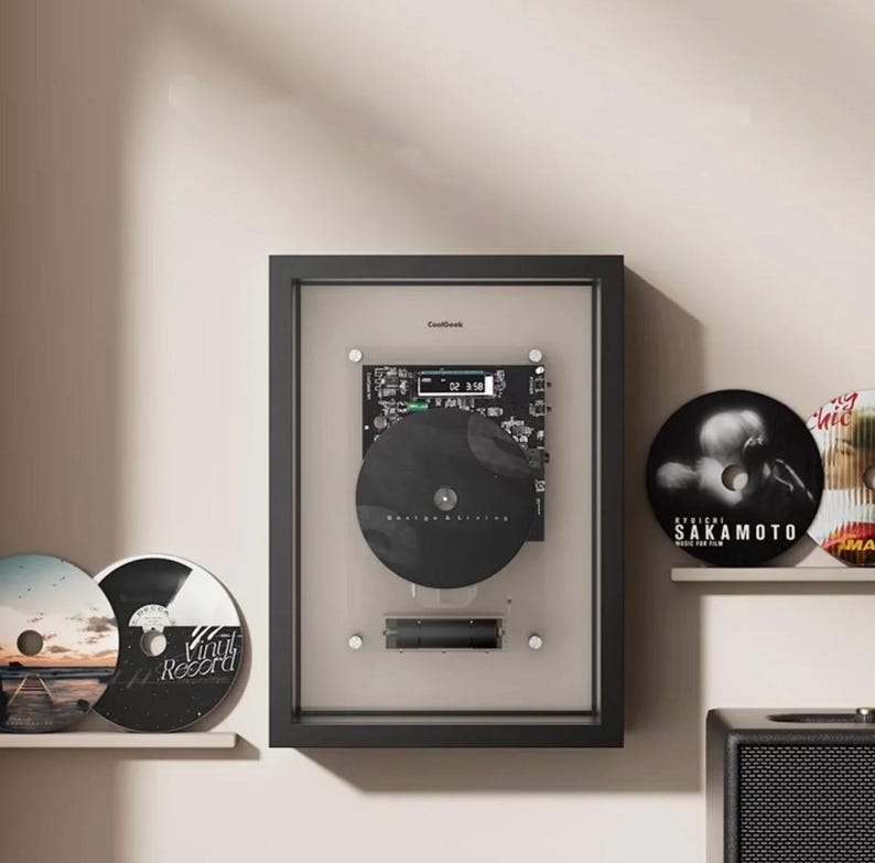 May include: A black framed vinyl record player with a black record and a digital display. Two vinyl records are on a shelf to the left, and two more are on a shelf to the right. The wall is a neutral colour.