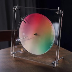 Może przedstawiać: Nowoczesny, minimalistyczny zegar z przezroczystą akrylową ramą. Tarcza zegara ma gradient kolorów od czerwonego do zielonego. Srebrne metalowe pręty i okucia mocują tarczę w ramie. Zegar stoi na drewnianej powierzchni.