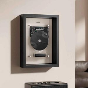 May include: A black framed CD display case mounted on a wall. The case has a black frame, a clear acrylic front, and a black CD. The text "CoolGreek" is visible at the top. A black speaker is at the bottom.