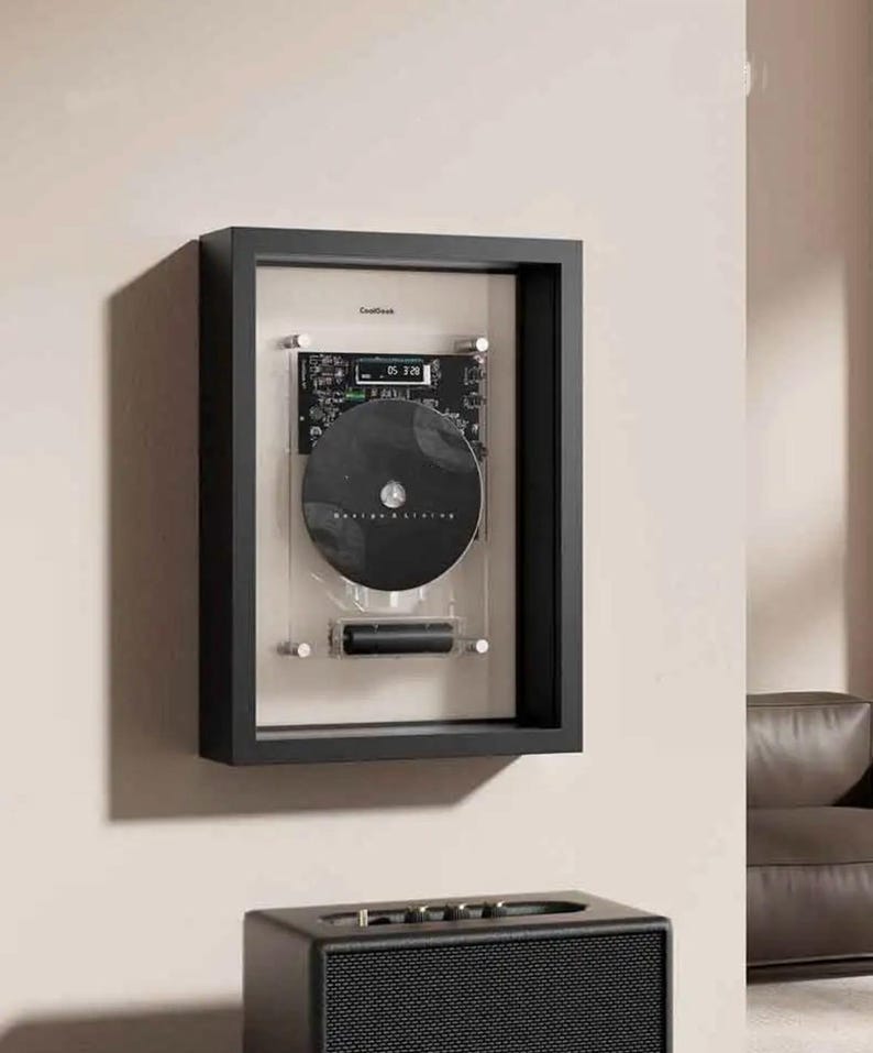 May include: A black framed CD display case mounted on a wall. The case has a black frame, a clear acrylic front, and a black CD. The text "CoolGreek" is visible at the top. A black speaker is at the bottom.