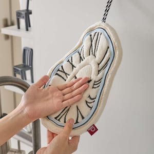 May include: A cream-colored, melting clock-shaped hand towel with black Roman numerals and clock hands. The towel has a light blue border and a black and white striped hanging loop. A red tag is attached to the towel.
