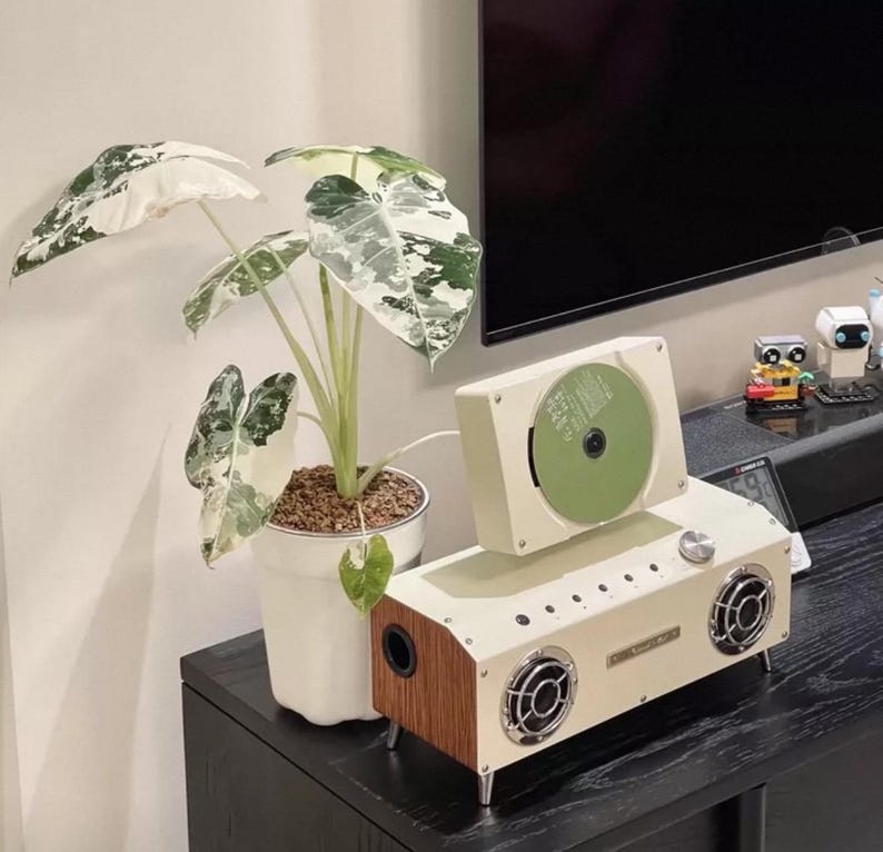 Puede incluir: Un reproductor de CD port&aacute;til de color crema con paneles laterales de imitaci&oacute;n madera y dos altavoces frontales, con una ranura vertical para discos con un CD verde insertado. Junto a &eacute;l, una maceta de cer&aacute;mica blanca contiene una planta Alocasia de hojas variegadas.