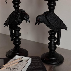 Pode incluir: Dois castiçais pretos com tema de corvo. Cada suporte apresenta um corvo empoleirado em uma base decorativa. Os corvos têm bicos abertos e olhos vermelhos. Uma caixa de fósforos e um livro estão em primeiro plano.