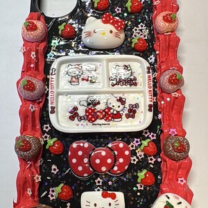 May include: A decorative phone case featuring Hello Kitty and strawberry-themed embellishments. The case has a black background with glitter and star accents, a red border, and various 3D elements, including a Hello Kitty face, a plate with Hello Kitty images, and strawberry-shaped decorations.