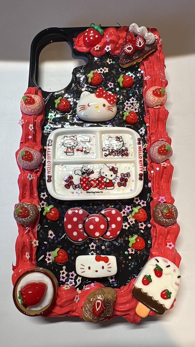 May include: A decorative phone case featuring Hello Kitty and strawberry-themed embellishments. The case has a black background with glitter and star accents, a red border, and various 3D elements, including a Hello Kitty face, a plate with Hello Kitty images, and strawberry-shaped decorations.