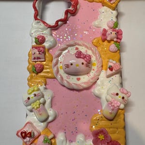 May include: A pink phone case decorated with Hello Kitty, strawberries, flowers, and dessert-themed embellishments. The case has a glittery background and a wavy border. The design includes various 3D elements.