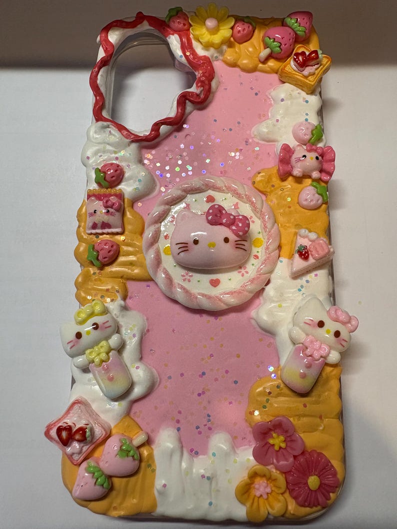May include: A pink phone case decorated with Hello Kitty, strawberries, flowers, and dessert-themed embellishments. The case has a glittery background and a wavy border. The design includes various 3D elements.