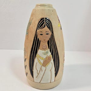 Puede incluir: Un jarrón de cerámica beige con una ilustración en relieve de una persona con cabello largo y negro, que viste una prenda blanca y un collar de cuentas amarillas, sosteniendo una pequeña flor. La superficie tiene un acabado texturizado con motivos florales.