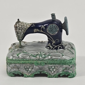 Puede incluir: Un joyero decorativo con forma de máquina de coser antigua. El brazo superior es azul oscuro con filigrana plateada y pedrería transparente. La base es una caja rectangular con acabado gris jaspeado, bordes verdes y detalles de pedrería.