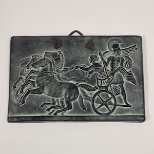 Puede incluir: Una placa de pared rectangular de color verde oscuro y negro con un relieve de un carro tirado por tres caballos. Una figura con casco está de pie en el carro sosteniendo las riendas, mientras otra figura está a su lado. Un cordón de cuero está unido.