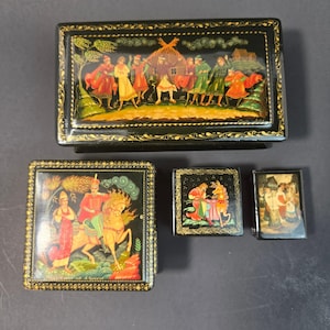 Puede incluir: Cuatro cajas de laca negra con escenas pintadas a mano. La caja rectangular más grande muestra figuras bailando en un paisaje. Una caja cuadrada presenta a una persona a caballo. Dos cajas más pequeñas representan una figura sentada y un grupo.